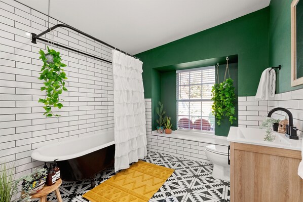 A bathroom with a white bathtub, a wooden vanity, a window with a view, and green plants.