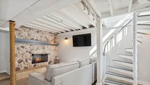 A modern living room with a stone fireplace, a grey sofa, a TV mounted on the wall, and a white spiral staircase.