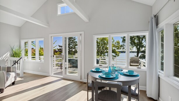A bright, open-plan living area with a dining table set for two, large windows offering a view of the outdoors, and a staircase leading to an upper level.