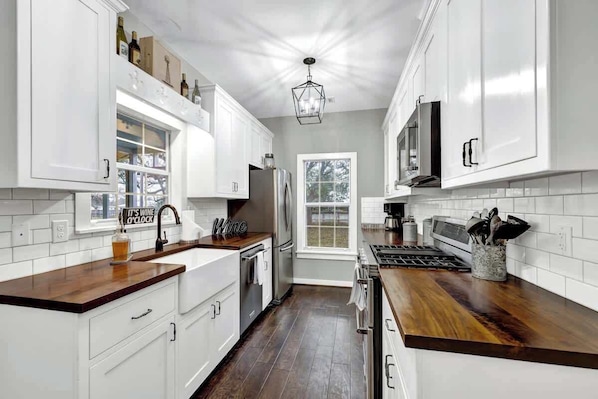 A modern kitchen with white cabinets, a wooden countertop, stainless steel appliances, and a window with a view of trees.