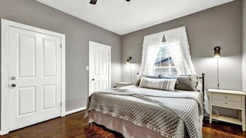 Une chambre à coucher avec un lit, des tables de chevet, un ventilateur de plafond et une fenêtre avec un rideau blanc.