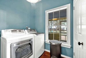 A laundry room with a front-loading washer and dryer, a window with a view of the outdoors, and a trash can.