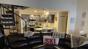 En moderne stue med en brun lædersofa, et køkken i baggrunden og en trappe, der fører til en etage ovenpå.