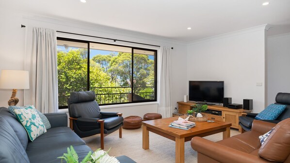Uma sala de estar moderna com uma janela ampla, uma TV de tela plana, sofás de couro, uma mesa de centro de madeira e uma mesa lateral com abajur.