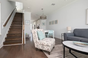 Un salón amplio con escalera, un sofá gris, un sillón estampado y una zona de comedor con mesa y sillas blancas.