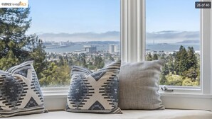 Un siège de fenêtre agrémenté de trois coussins à motifs et offrant une vue sur le paysage urbain et les montagnes.