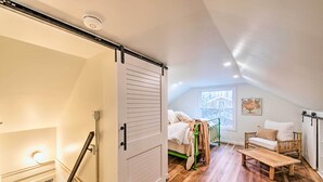 A cozy attic bedroom with a loft bed, a green chair, a wooden bench, and a window with a view of trees. - Bloomington Bungalow - Steps to Campus! (Bloomington)
