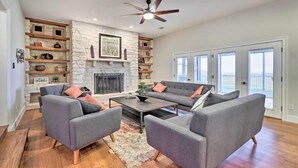 A living room with a stone fireplace, grey sofas, wooden shelves, and a ceiling fan. - Modern Lakehouse w/ Stunning Views & Hot Tub! (Granger)