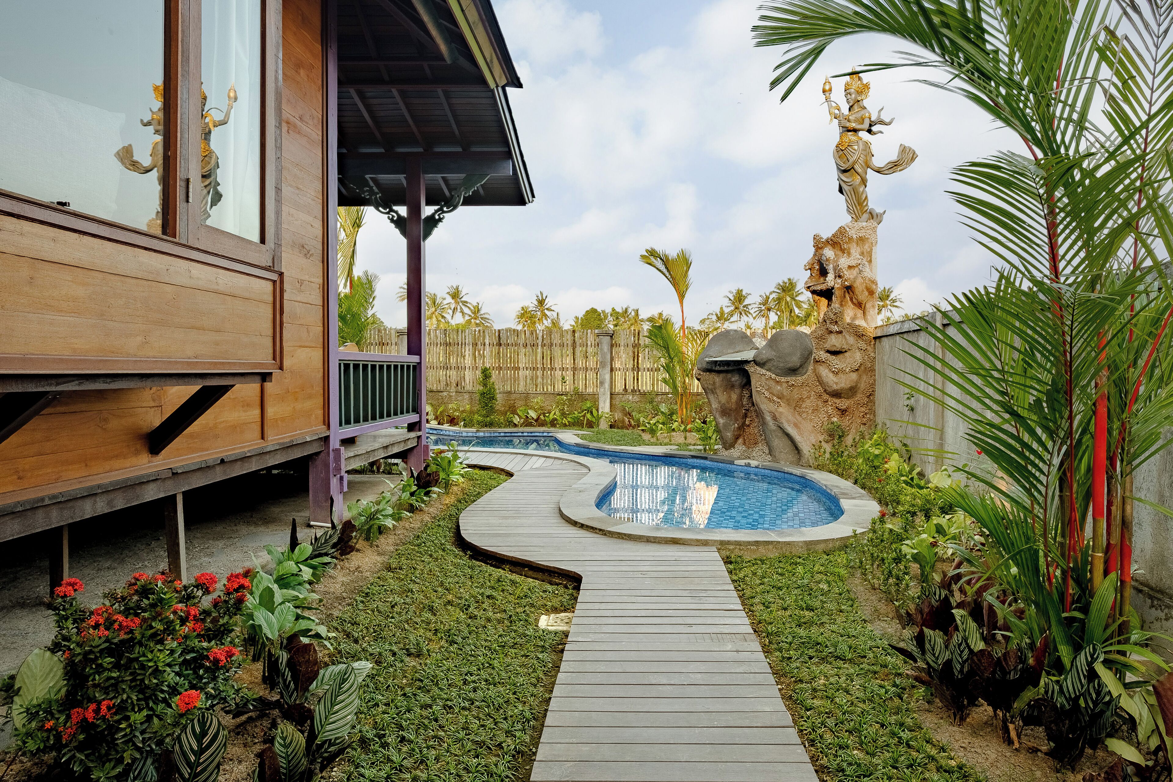 Un terrazzo in legno che conduce a una piscina a forma di curva, anch'essa circondata da un terrazzo in legno, tra piante tropicali e una statua decorativa.
