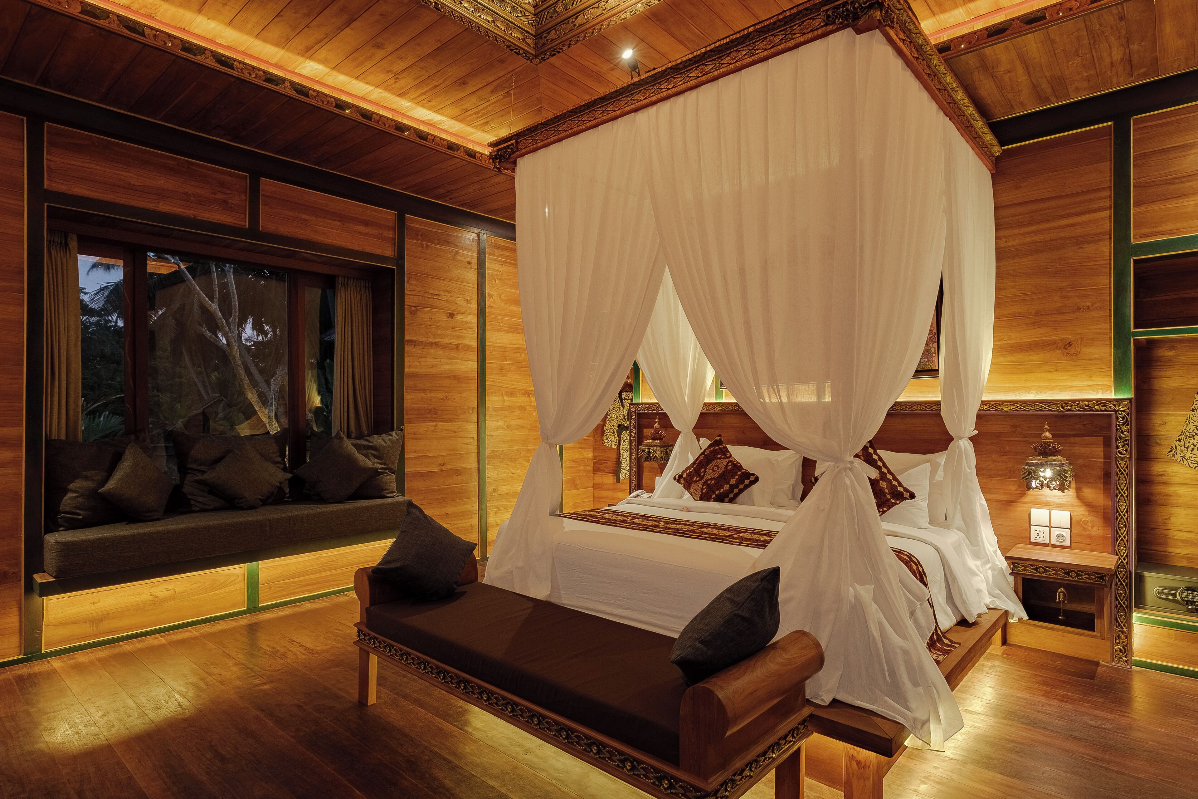 A four-poster canopy bed with white linens and a wooden headboard, a bench with cushions, and a nightstand with a lamp.