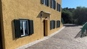 Un bâtiment à l’extérieur recouvert d’un enduit jaune, doté de nombreuses fenêtres avec volets, d’une entrée pavée de pierres et d’un portail en métal vert.