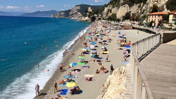 Une plage bondée où les gens se prélassent au soleil et se baignent, une promenade en bois et des bâtiments situés sur une colline.