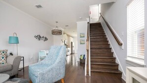 Un vaste salon agrémenté d’un escalier en bois, d’un fauteuil bleu et offrant, au fond, une vue sur la cuisine.