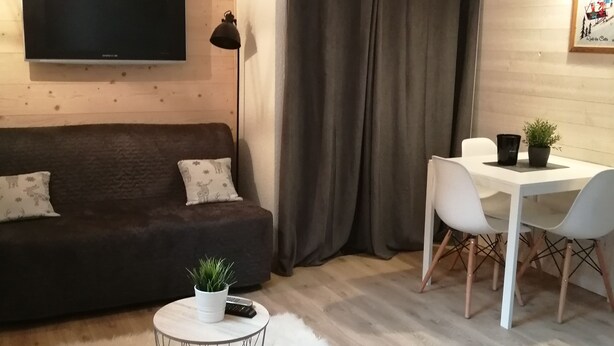 A cozy living room with a dark sofa, a white round coffee table with a plant, a white dining table with chairs, and a flat-screen TV mounted on the wall. - Studio in the heart of nature, 800 m from La bresse-hohneck ski resort (La Bresse)