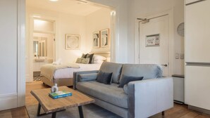 Ett kompakt hotellrum med en säng, en grå soffa, ett litet träbord och ett litet kök.