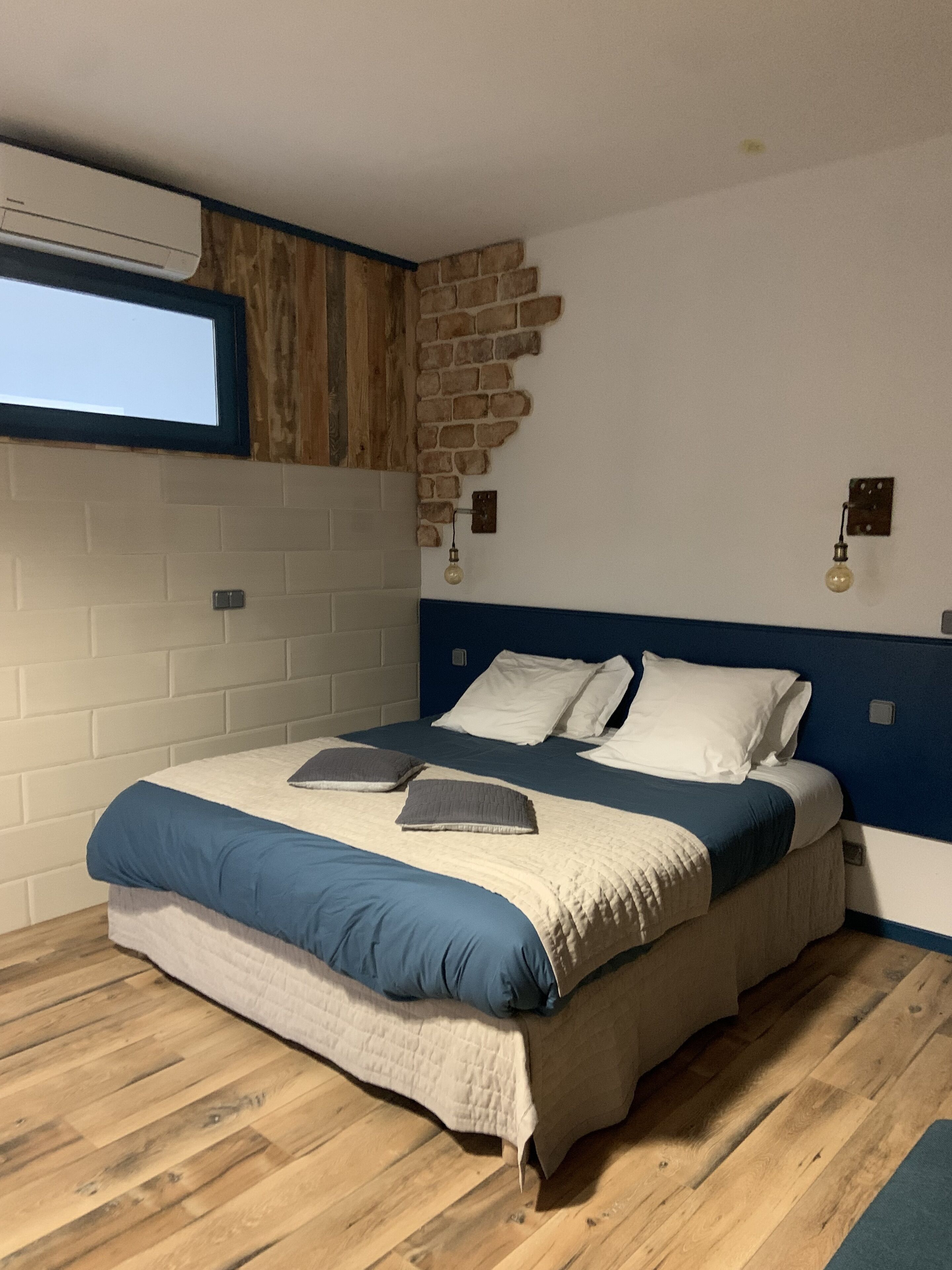 A bedroom with a bed, wooden flooring, a brick wall, and a window.