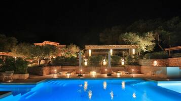 Un área de piscina iluminada de noche con sillones de descanso y una pérgola.