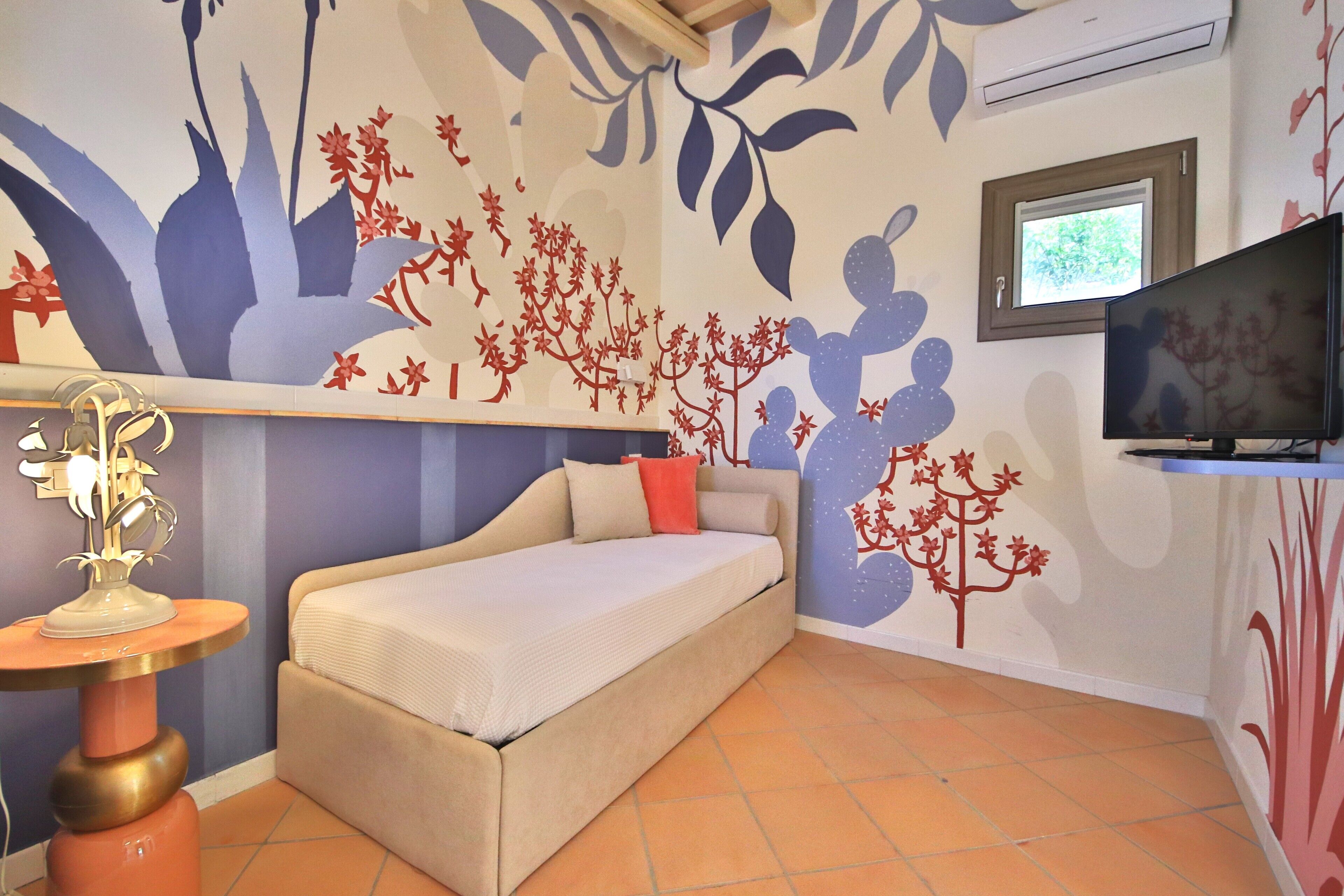 A bedroom with a bed, a TV, and a wall with floral and tree designs.