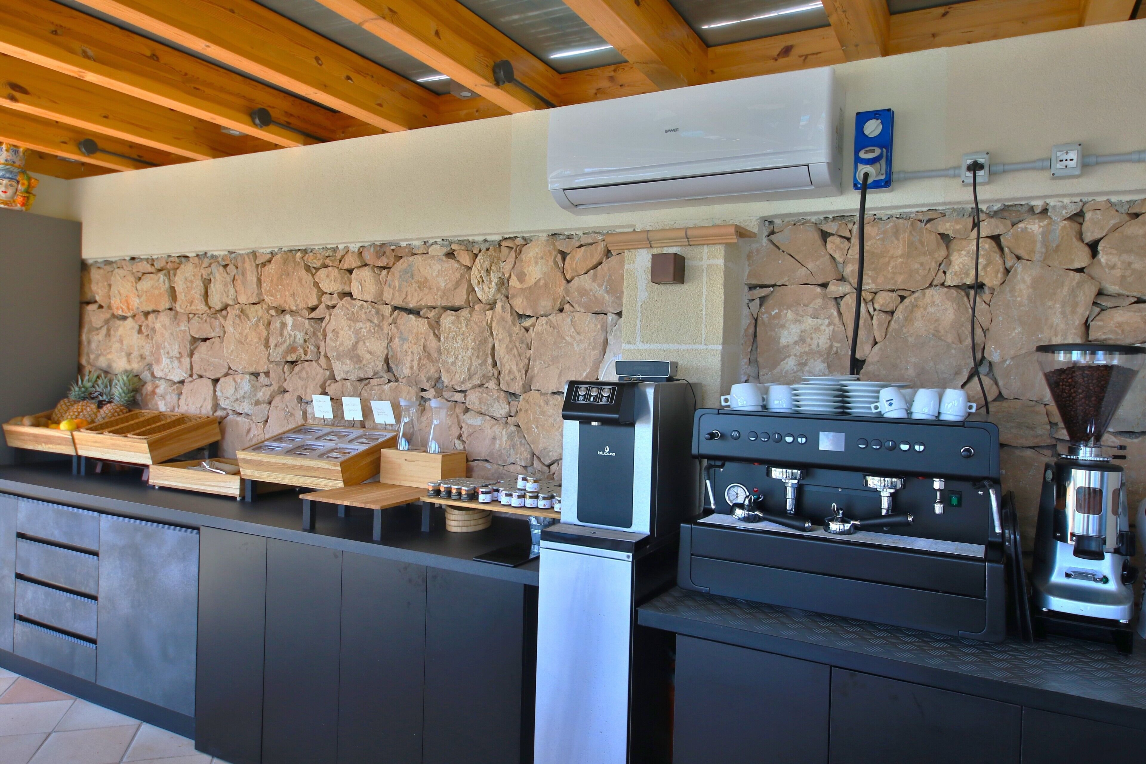 A kitchen area with a stone wall, coffee machines, and a fruit display.