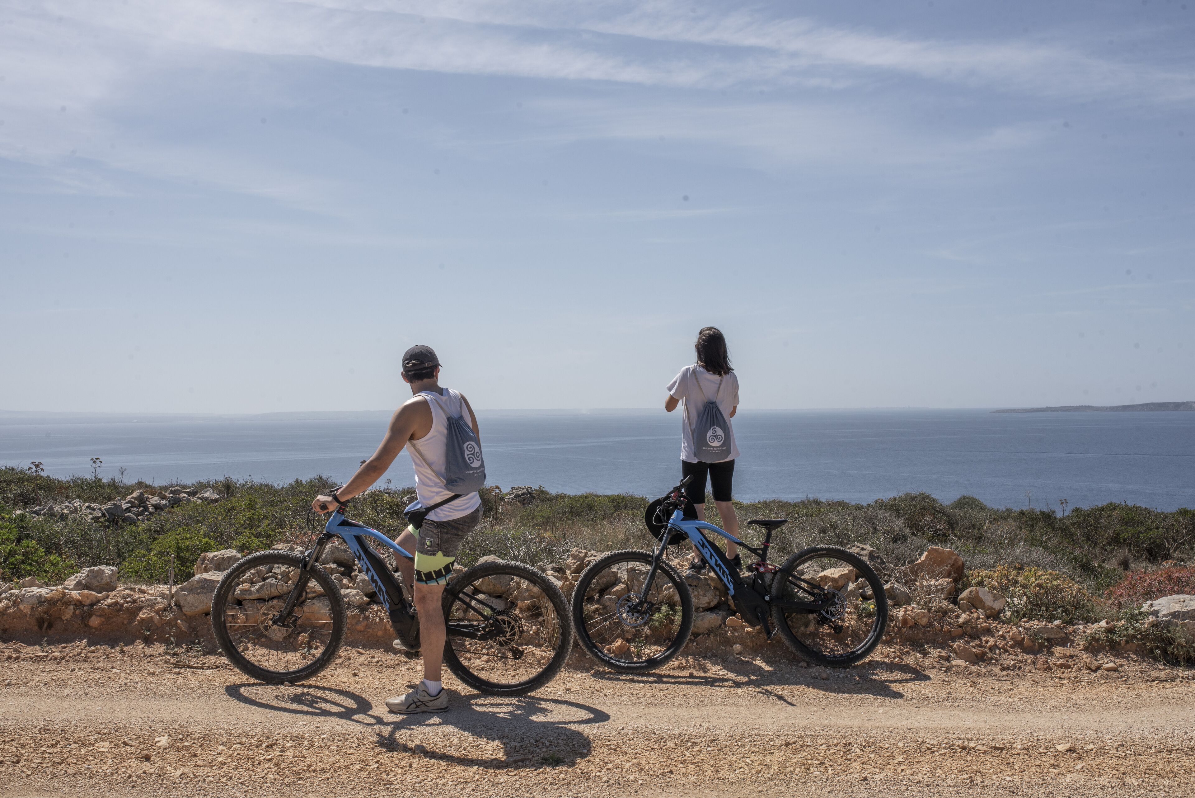 Two people on a dirt path with bicycles, overlooking a coastal landscape.