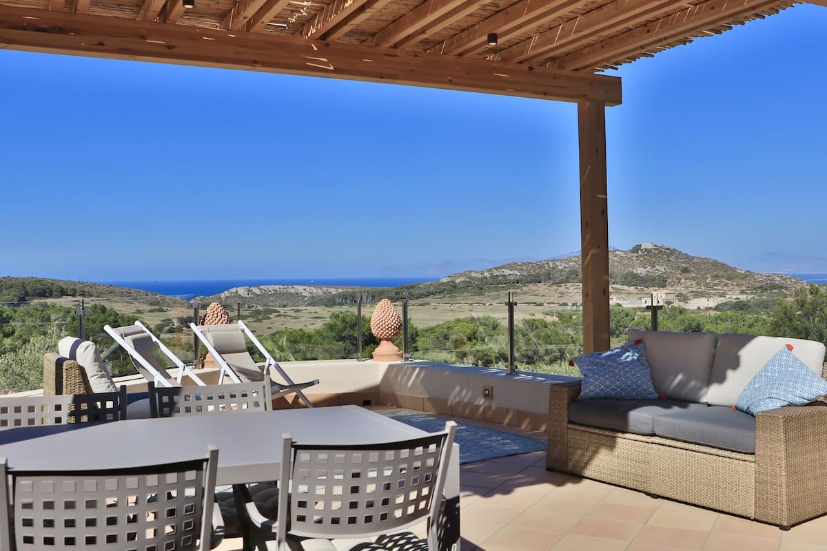 Eine Terrasse im Freien mit Tisch, Stühlen und einer Couch mit Kissen, mit Blick auf eine malerische Landschaft mit Hügeln und dem Meer.