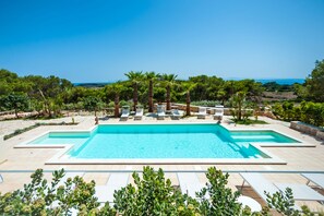 Une piscine rectangulaire à l’eau d’un bleu limpide, entourée de chaises longues et de palmiers, avec une vue sur la mer en arrière-plan.