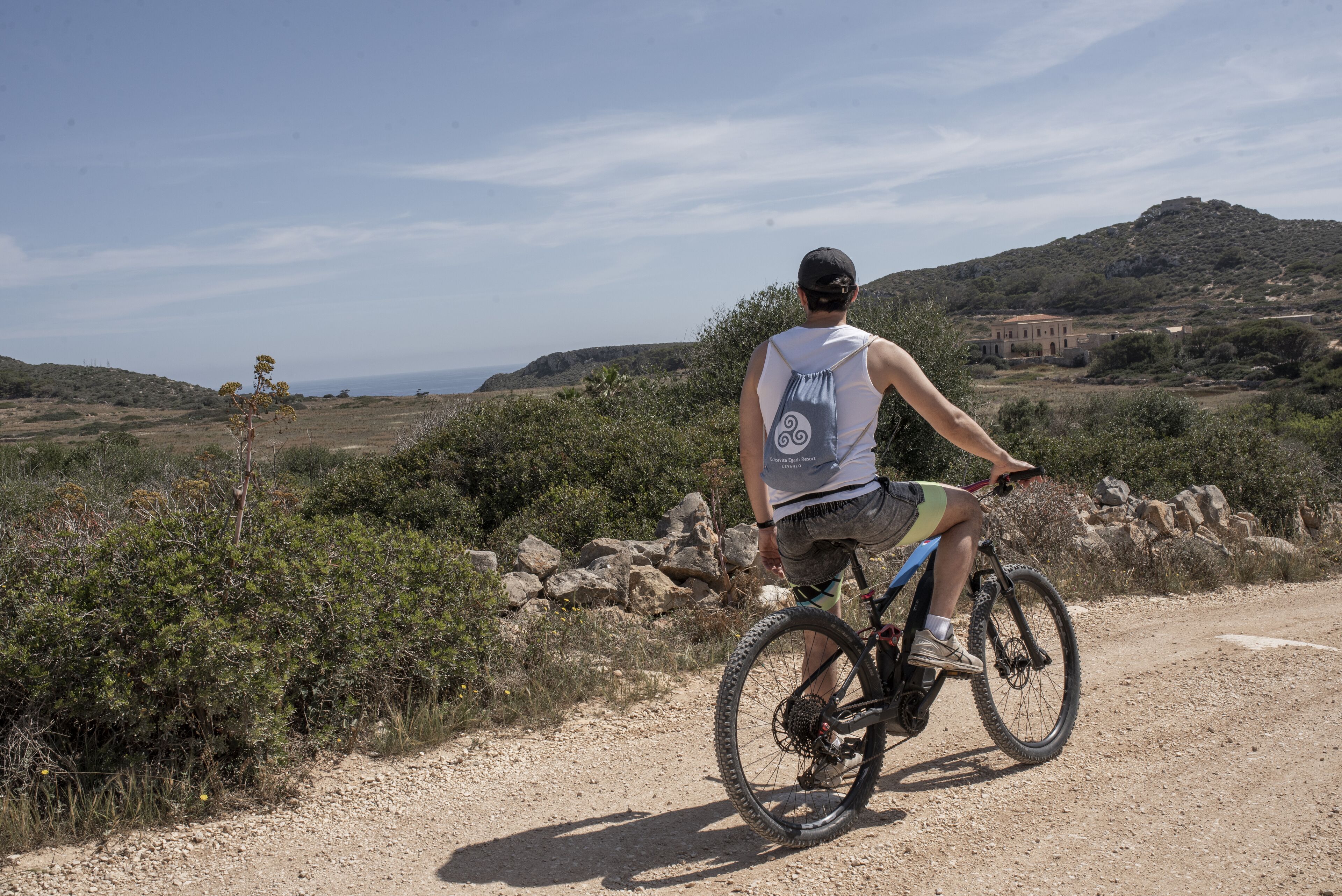 Una persona montando una bicicleta de montaña por un sendero de tierra con vista al mar y colinas al fondo.