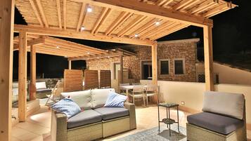 Un patio al aire libre con un pérgola de madera, un muro de piedra, una mesa de comedor con sillas y un sofá con cojines.