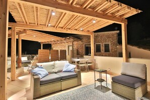 Un espace extérieur aménagé en terrasse, avec une pergola en bois, un mur en pierre, une table à manger avec des chaises et un canapé avec des coussins.