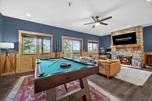 A pool table with a fire pit and a television in a room with large windows and a ceiling fan.