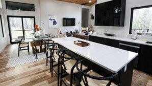 A modern kitchen with a large island, black cabinets, and a dining area with chairs and a table. - Serene Retreat Awaits: Welcome to Endless Views,Your Luxurious Contemporary Home (Muskegon)
