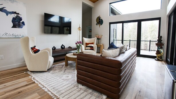 A spacious living room with a high ceiling, a large TV mounted on the wall, a comfortable sofa, a white armchair, and a coffee table. - Serene Retreat Awaits: Welcome to Endless Views,Your Luxurious Contemporary Home (Muskegon)