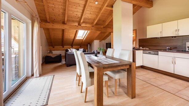 A spacious loft-style room with a wooden ceiling, a dining area with a wooden table and chairs, a kitchen with white cabinets, and a large window providing natural light. - Birkeneck 1 (DG), castle view dream holiday apartment (Schwangau)
