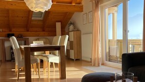 A cozy room with a wooden ceiling, a dining table with chairs, a sofa, and a large window with a view. - Birkeneck 1 (DG), castle view dream holiday apartment (Schwangau)