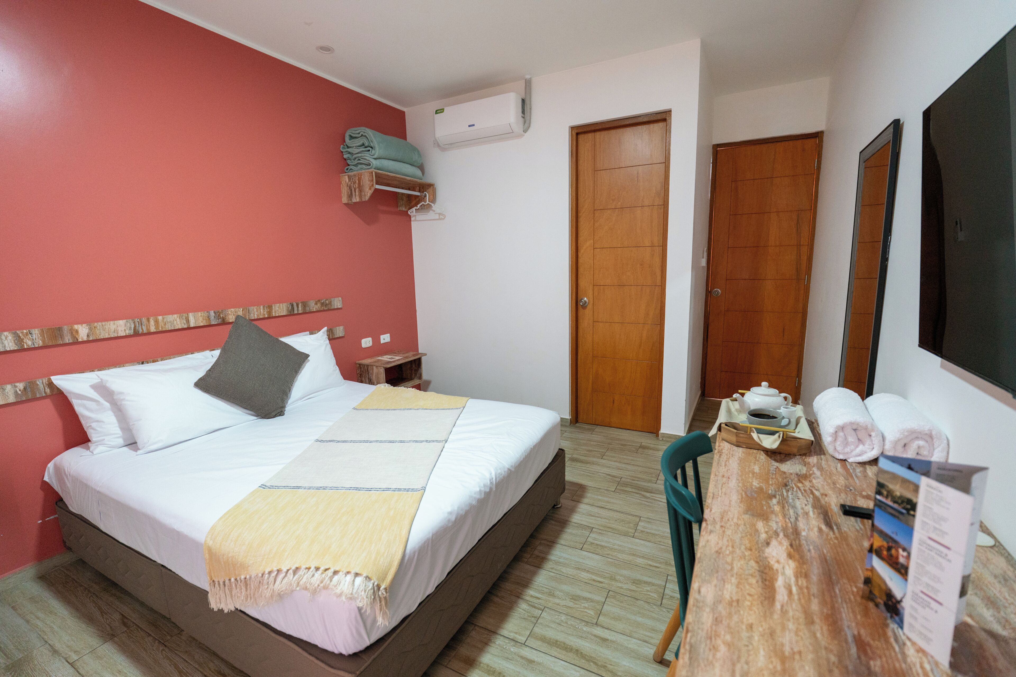 A hotel room with a bed, a wooden nightstand, a table with a chair, a wooden door, and an air conditioning unit.