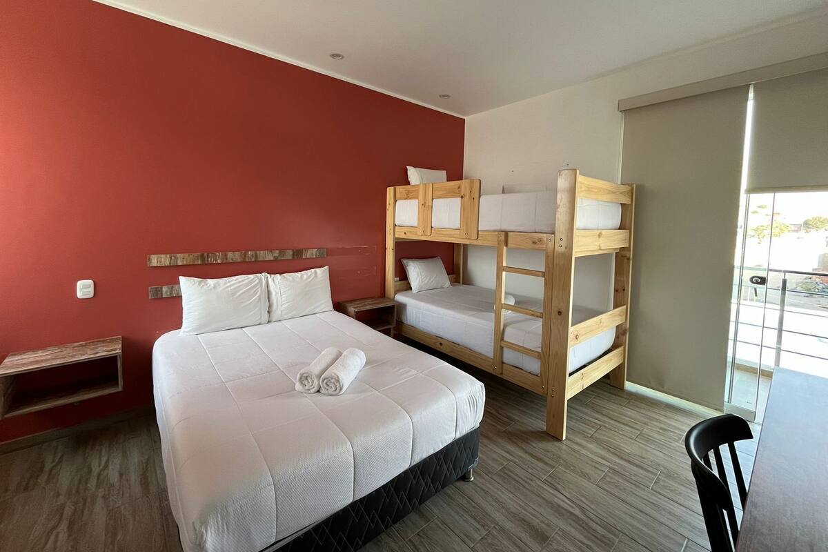 Une chambre d’hôtel avec un grand lit, deux lits superposés, une tête de lit en bois, une petite table de chevet et une chaise.