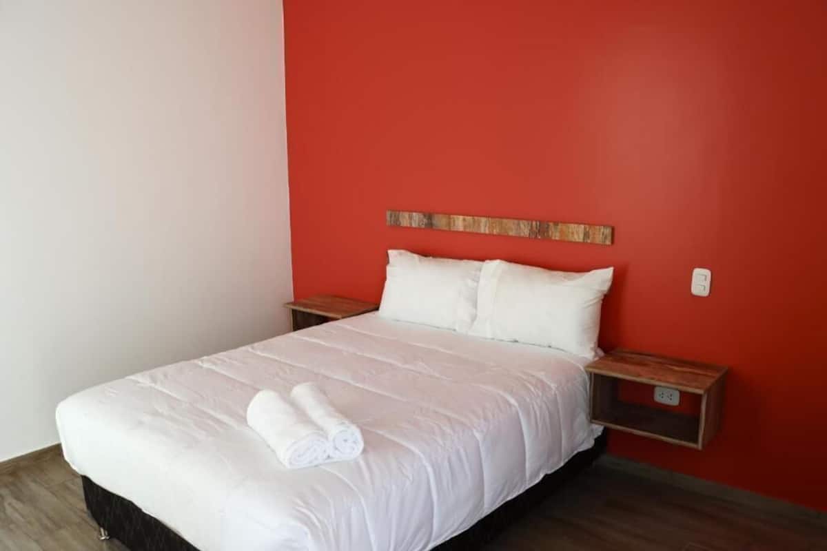 Un lit bien fait, avec du linge de lit blanc et une serviette enroulée, une table de chevet en bois et un mur rouge.