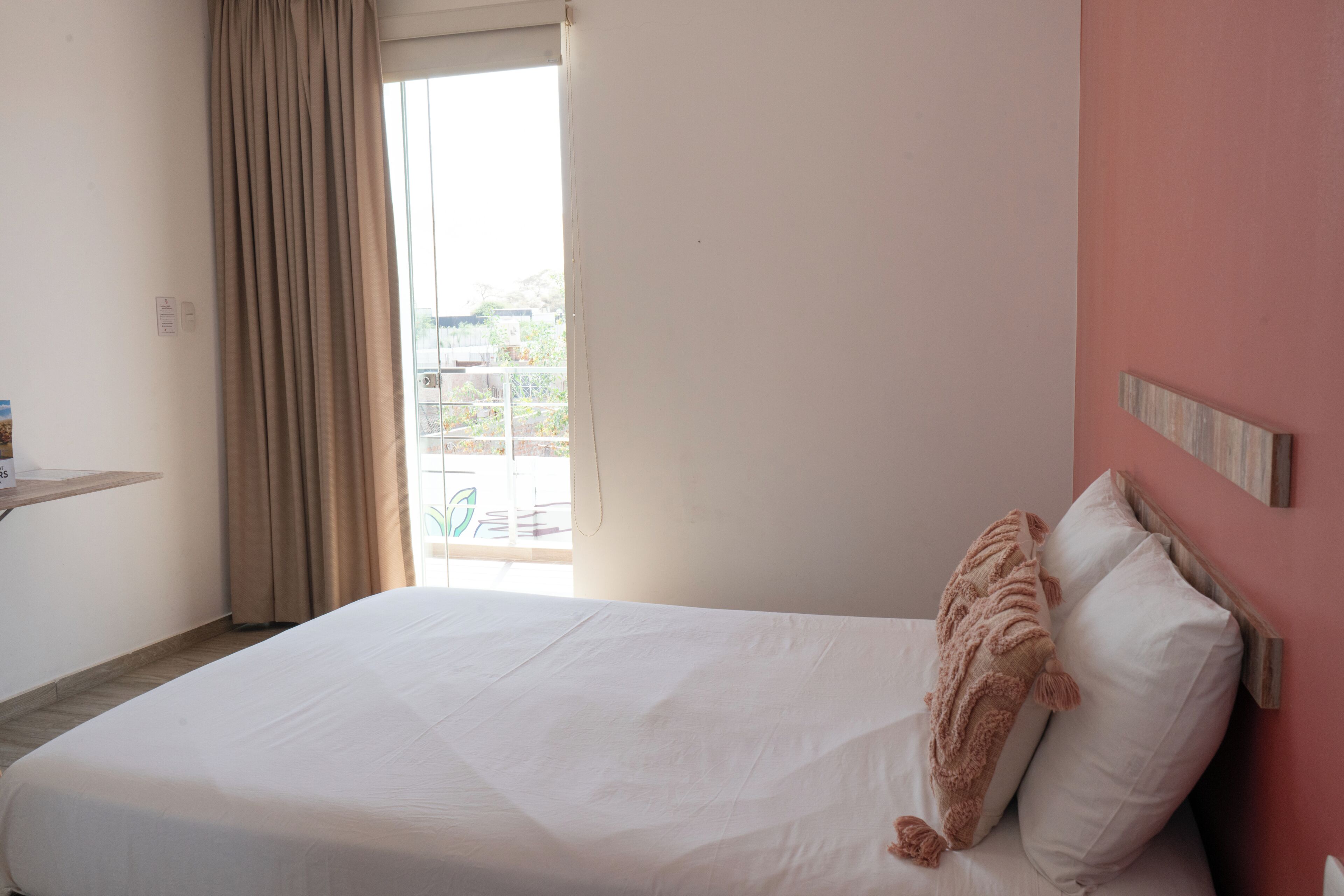 A hotel room with a bed, a window with curtains, a wooden headboard, and a wall-mounted shelf.