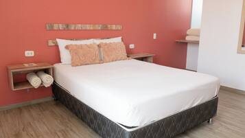 Una cama con colchón blanco y dos almohadas, una mesita de noche de madera con una toalla, y un estante en la pared con toallas.