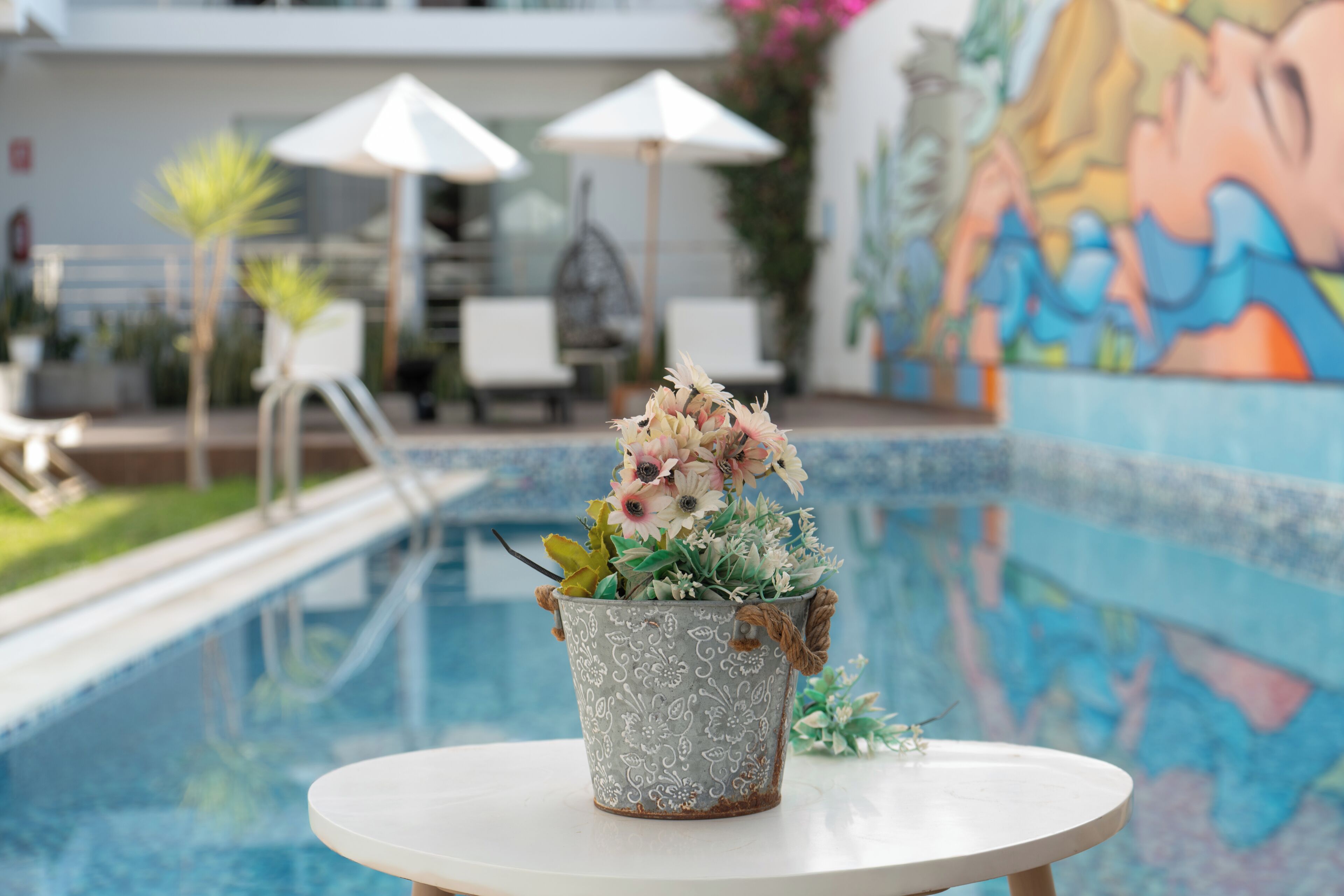 Zona de piscina con una maceta decorativa sobre una mesa, sillas de descanso, sombrillas y un mural.