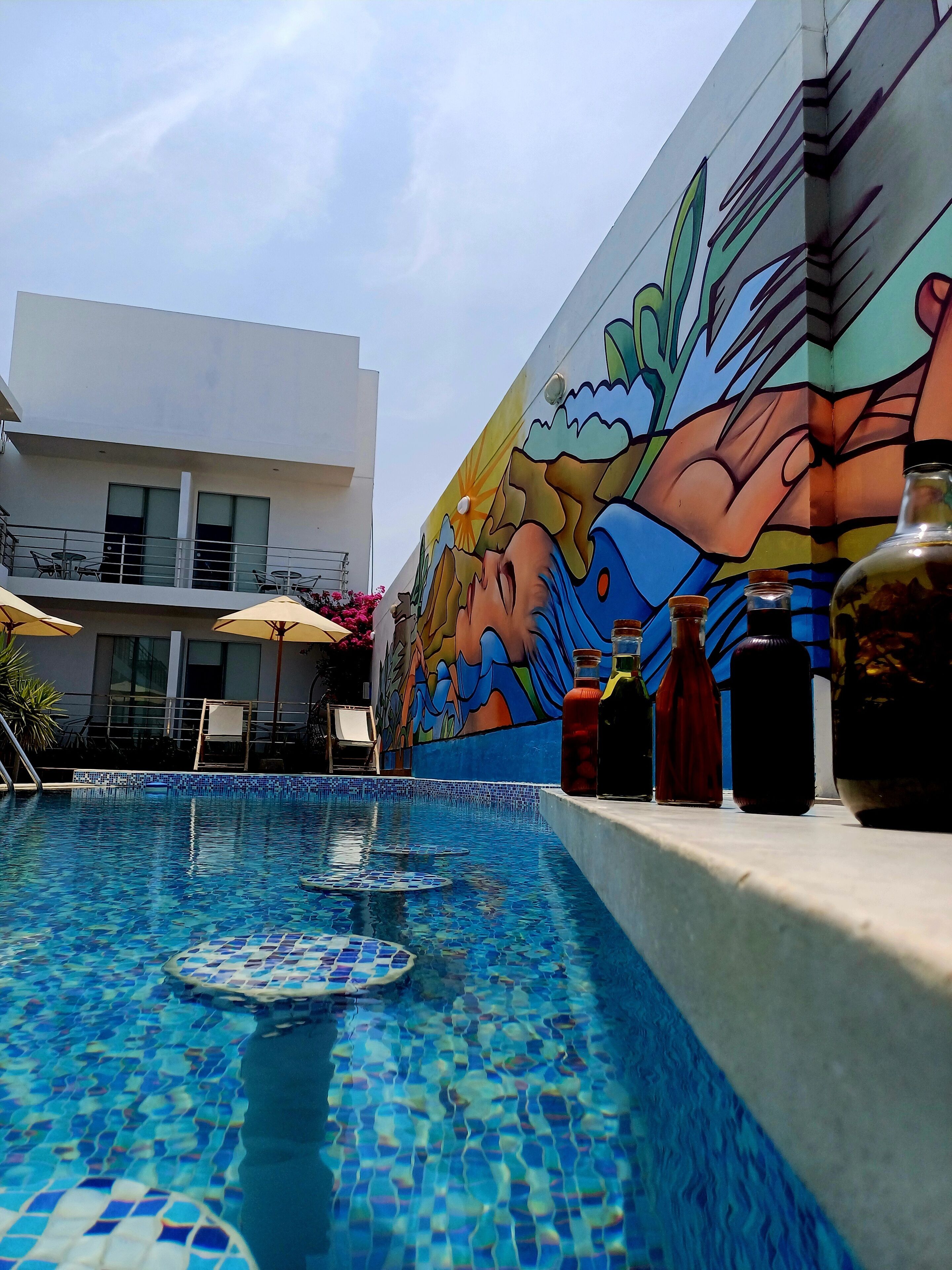 A swimming pool with mosaic tiles, a mural on the adjacent wall, and a few bottles on the poolside.