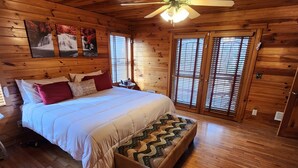 A wooden cabin bedroom with a bed, pillows, a nightstand, a ceiling fan, and a window with blinds.