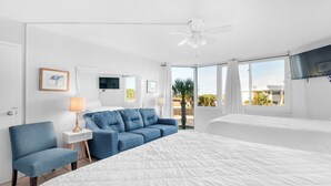A hotel room with a large bed, a blue armchair, a sofa, a TV, and a balcony with a view of palm trees.