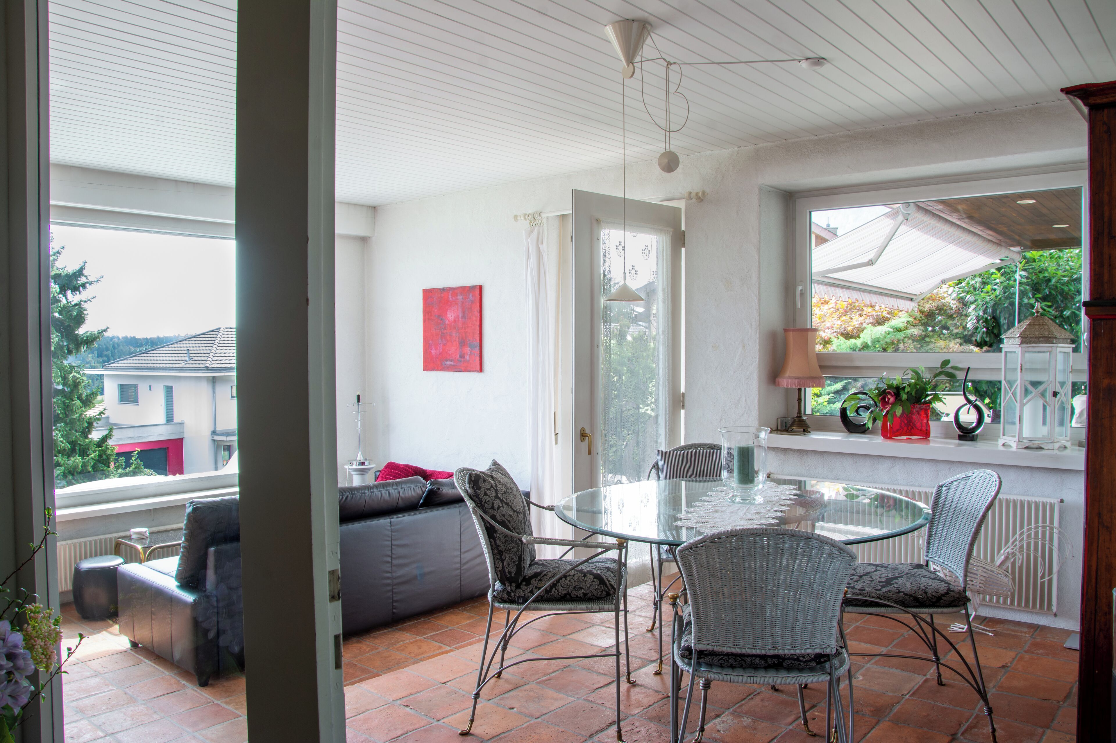 A dining area with a glass table and wicker chairs, a sofa with cushions, and a view of the outside through large windows.