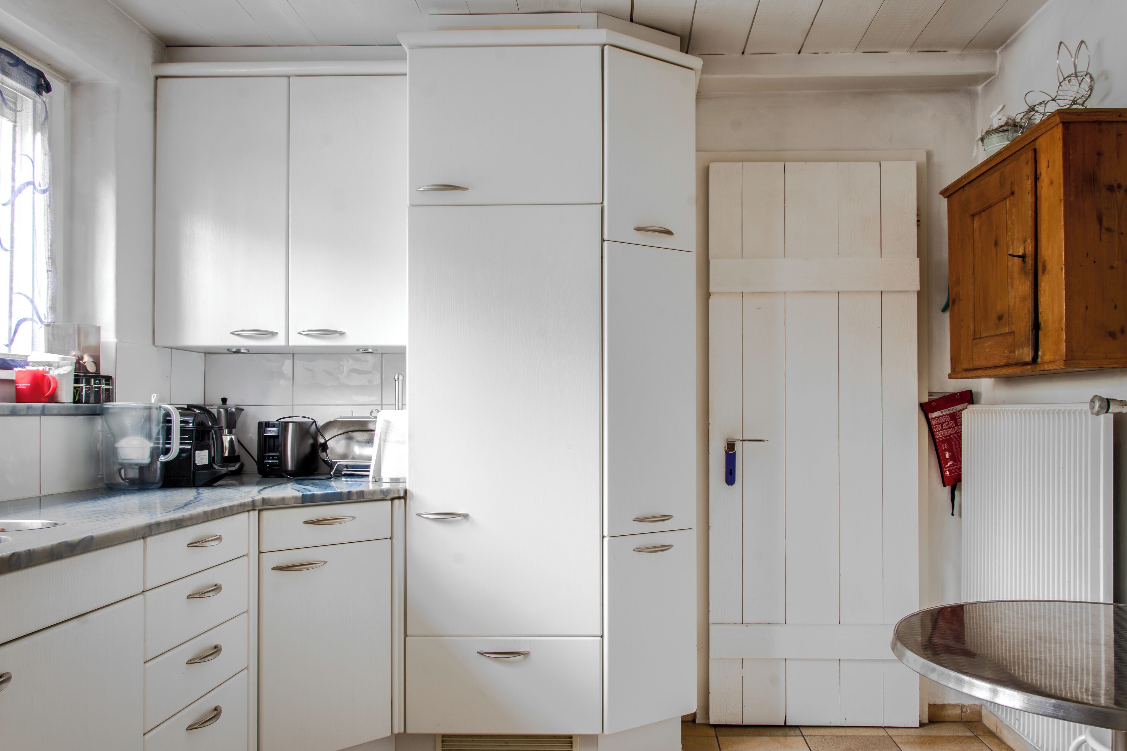 Een keuken met witte kastjes, een houten kast en een raam met gordijnen.