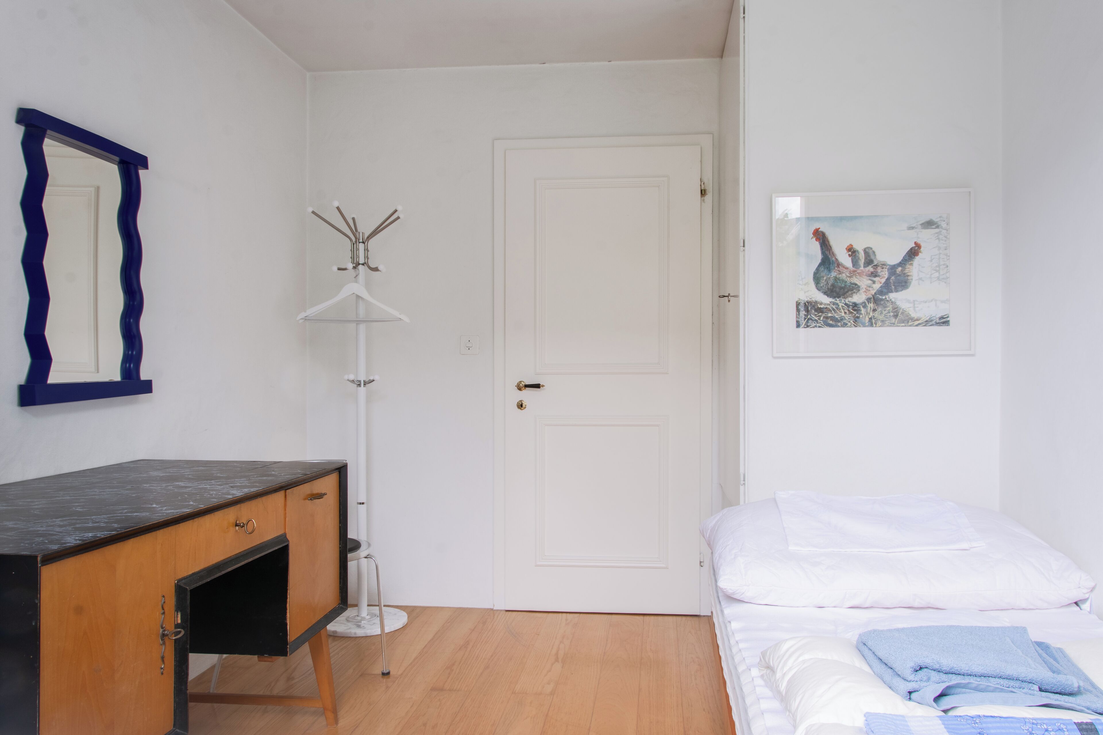 Ein Schlafzimmer mit einem Bett, einer Kommode, einer Tür und einem gerahmten Bild an der Wand.