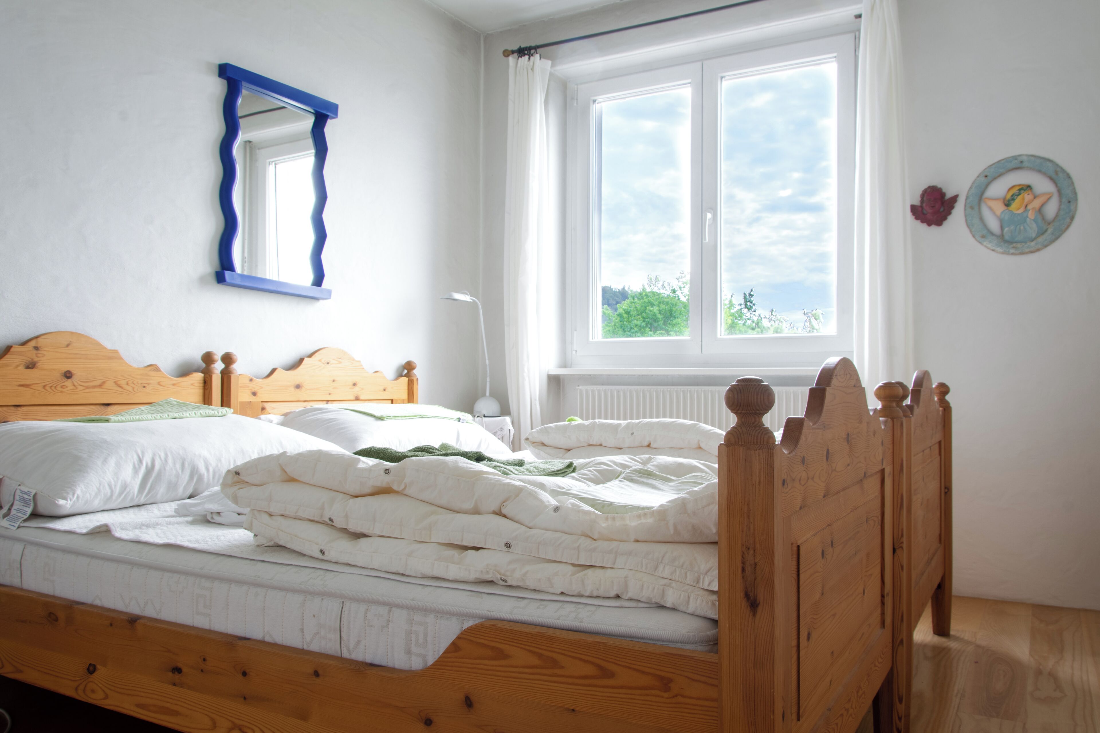 Un lit bien fait, avec une tête de lit en bois, une fenêtre ornée de rideaux blancs et un miroir à cadre bleu accroché au mur.