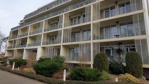 Un immeuble résidentiel à plusieurs étages, doté de balcons, d’un toit plat et d’un espace jardin agrémenté d’arbustes et d’un petit arbre.