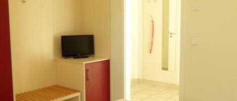Une chambre d’hôtel avec un lit, un téléviseur posé sur un meuble, une armoire rouge et une étagère en bois.
