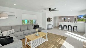 A modern living room with a grey sofa, a wooden coffee table, and a kitchen area with a bar and stools. - 5 mins to beach + Brand New Pool and Tiki Hut. (Oakland Park)